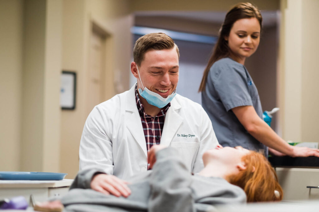 Dr. Dyer smiles while talking to teen patient during checkup while assistant looks on